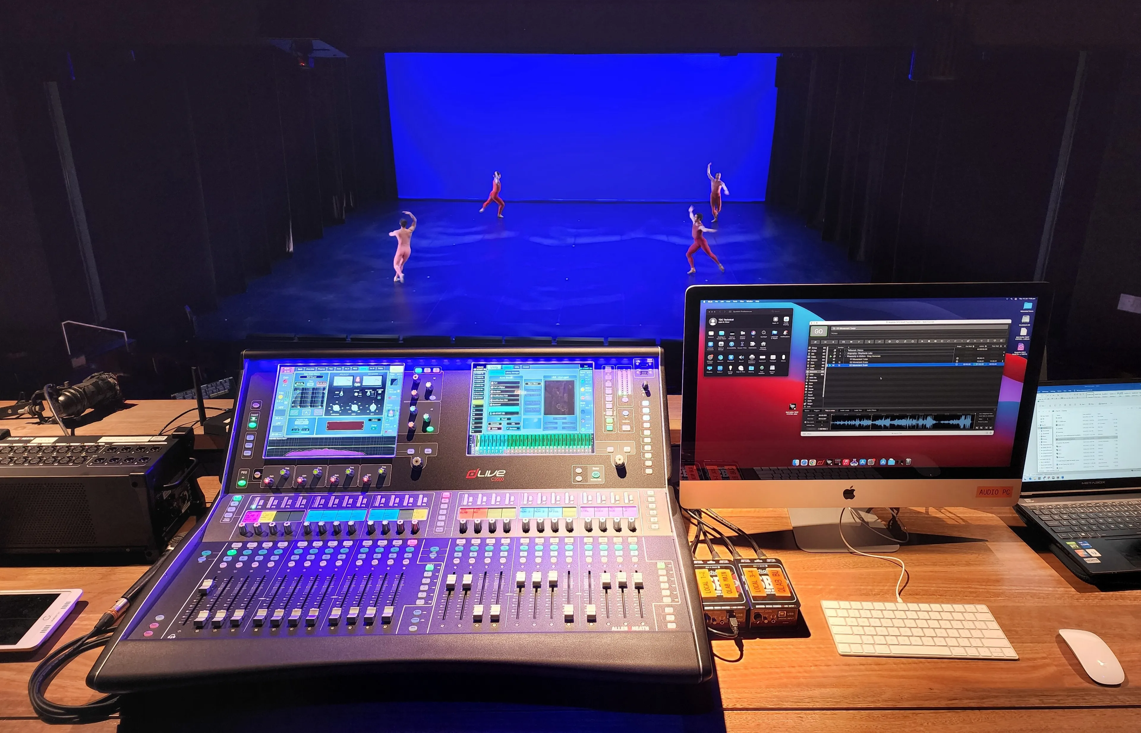 A mixing desk at front of house, dancers on a blue-lit stage beyond